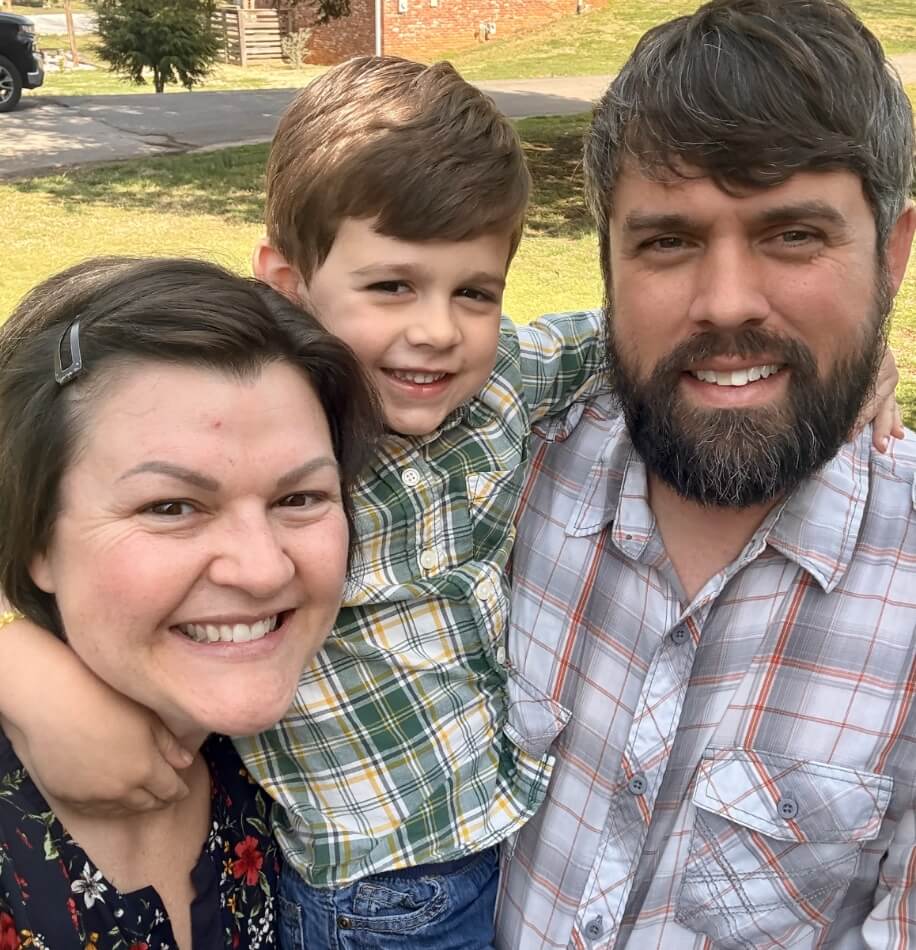 Two adults and a child smiling together outdoors with arms around each other.