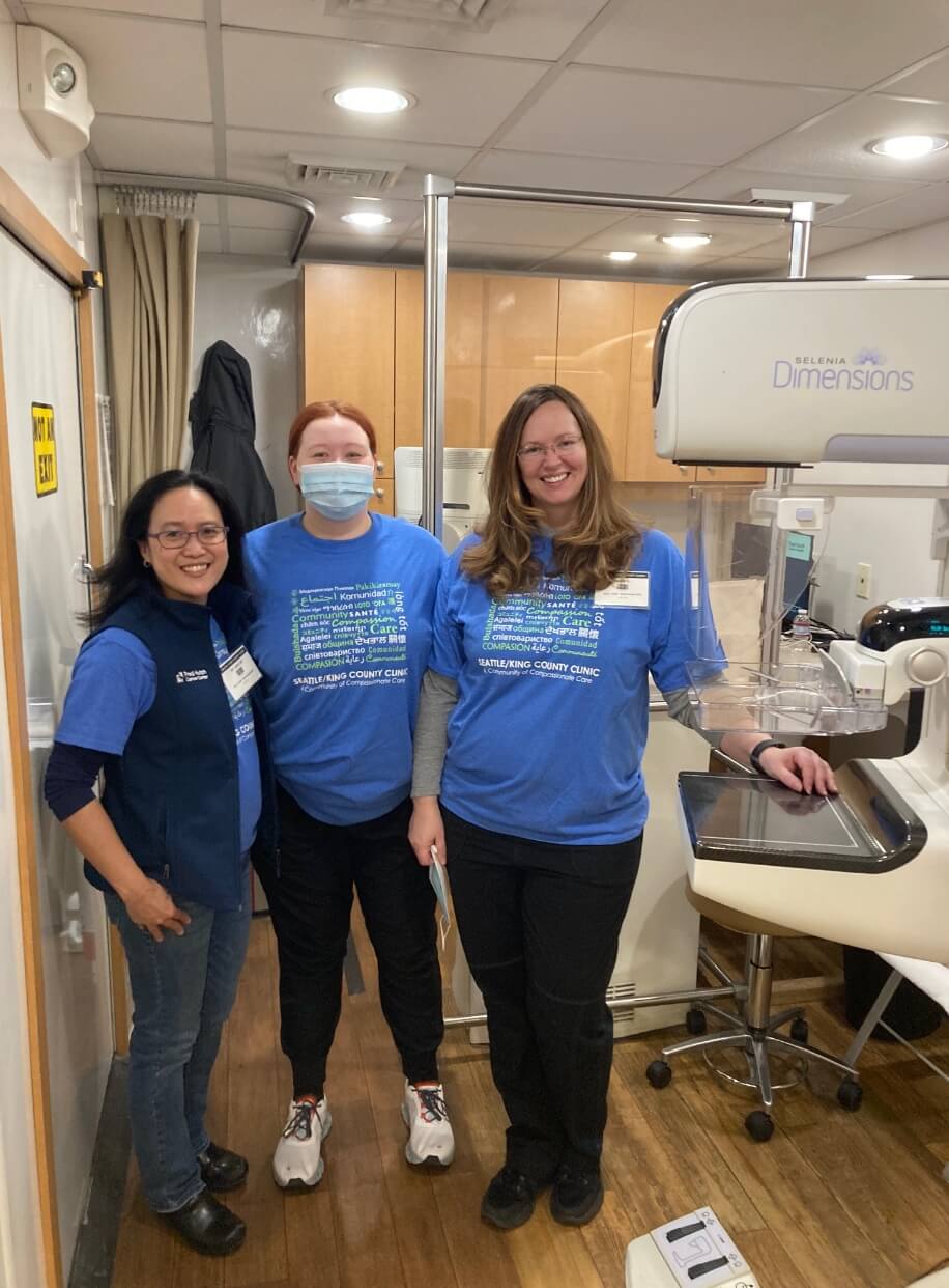 Three healthcare team members standing together in a clinic room beside mammography equipment.