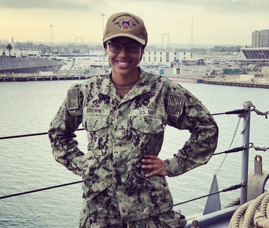 U.S. Navy sailor in camouflage uniform standing on the deck of a naval ship with harbor in the background.